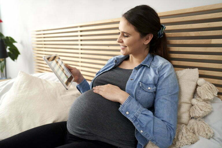 Caucasian pregnant woman sitting on bed, holding digital tablet